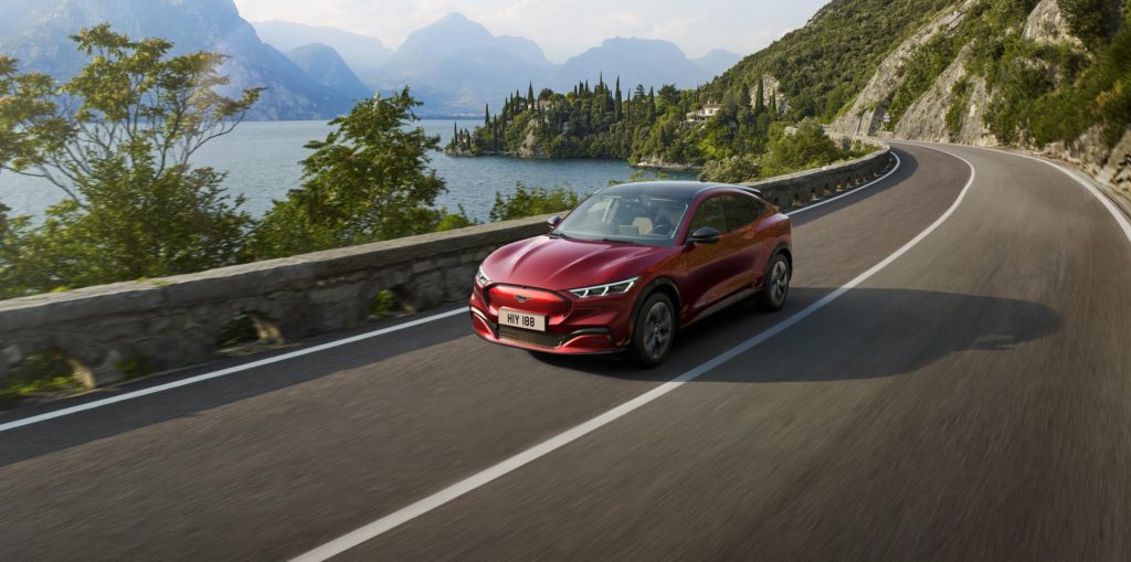 Ford Mustang Mach-E auf einer Straße neben einem See mit Bergen im Hintergrund